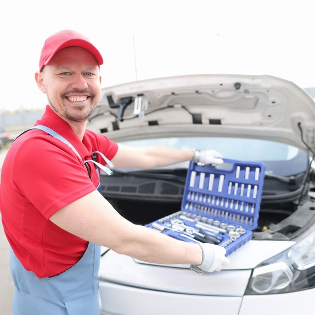 Photo: Smiling mechanic in a red cap and blue overalls with a socket set open on the engine of a white car