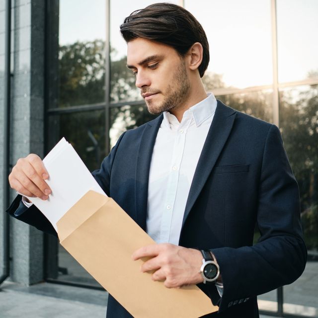 Photo: Man in a navy blazer taking a letter out of a brown envelope outside a glass office building