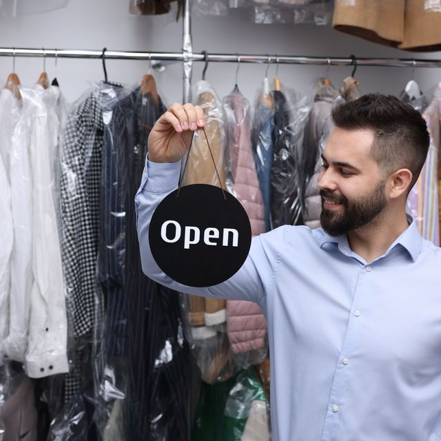 Photo: Smiling man holding an Open sign among racks of clothes in plastic covers at a dry cleaner