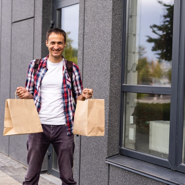 Photo: Smiling man in a plaid shirt carrying two brown paper grocery bags on a sidewalk outside a building
