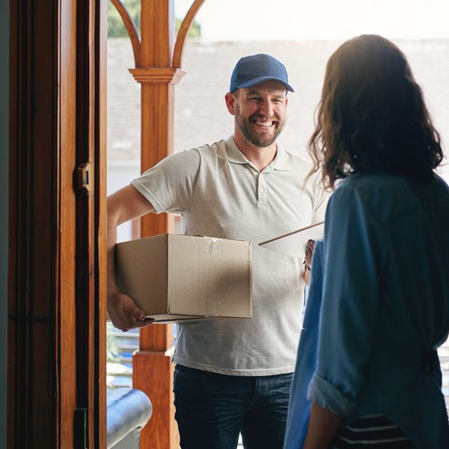 Photo: Smiling delivery man in a blue cap holding a cardboard box at a front door while a woman faces him
