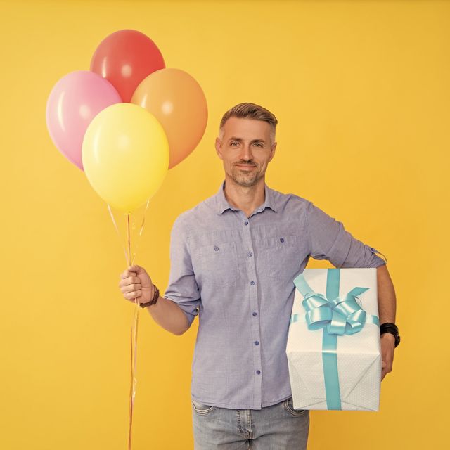 Photo: Man holding a bunch of balloons and a wrapped gift box against a yellow background