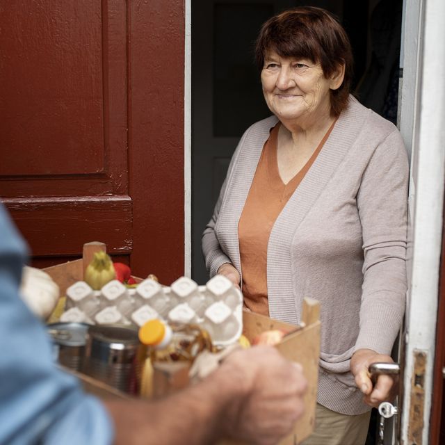 Photo: Smiling older woman at her open front door receiving a box of groceries from a person in a blue shirt