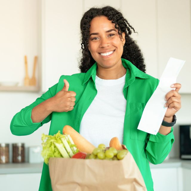 Photo: Smiling woman in a green shirt giving a thumbs up while holding a receipt and a paper bag of groceries in a bright kitchen