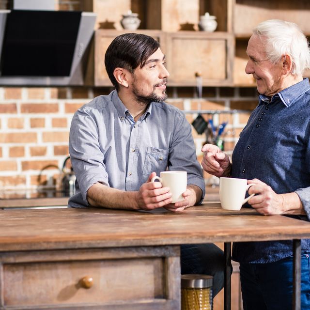 Photo: Two men laughing over coffee at a kitchen table, one younger and one older