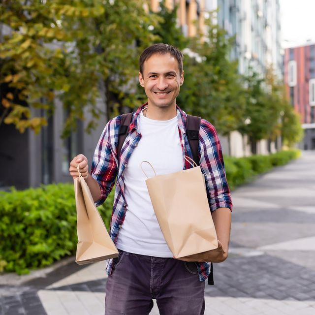 Photo: Smiling man with a backpack carrying two brown paper takeout bags along a tree-lined city path