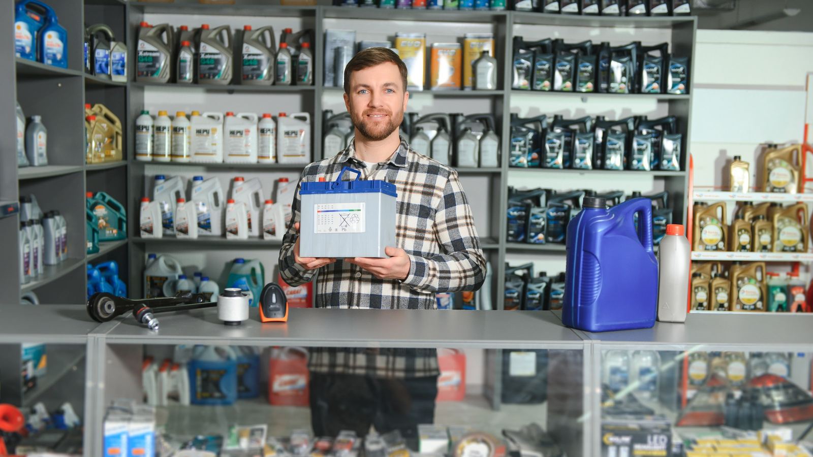 Smiling man in a plaid shirt holding a battery box behind the counter of an auto parts store with shelves of motor oil