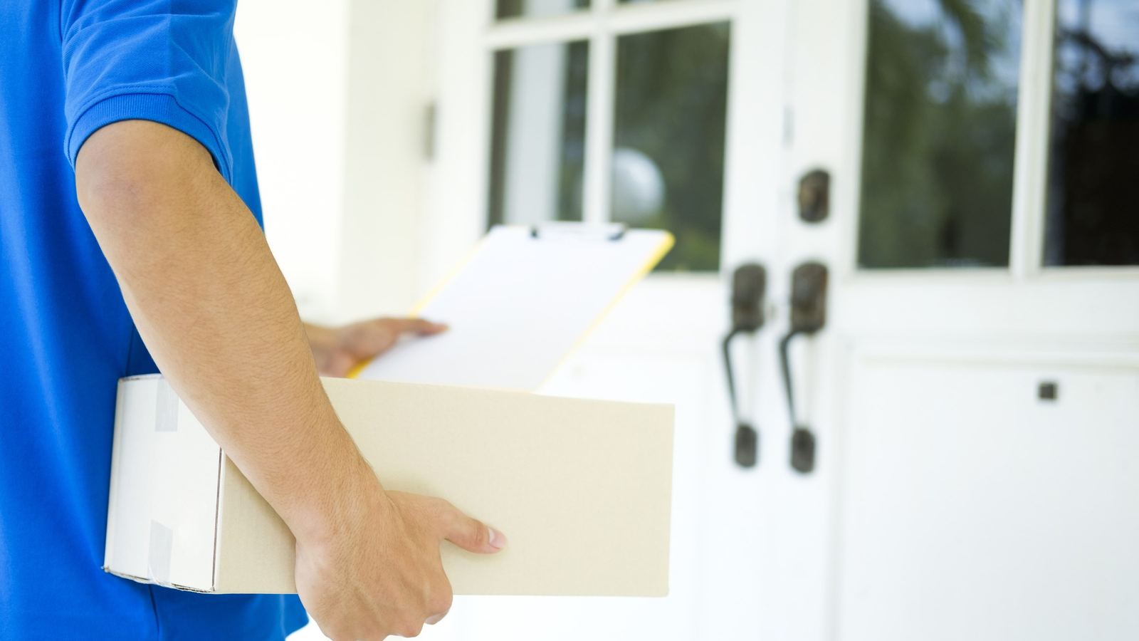 Delivery person in a blue shirt holding a cardboard package and a clipboard at a white front door