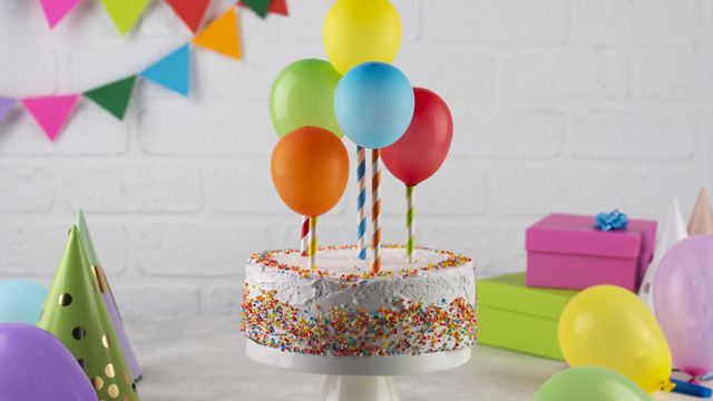 Birthday cake with colorful balloons, party hats and wrapped gifts on a table against a white brick wall
