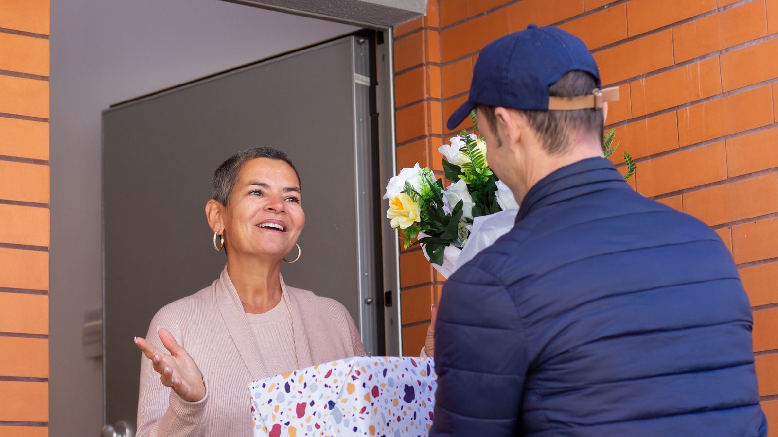 Delivery man in a blue cap handing a wrapped gift box to a smiling woman at her front door