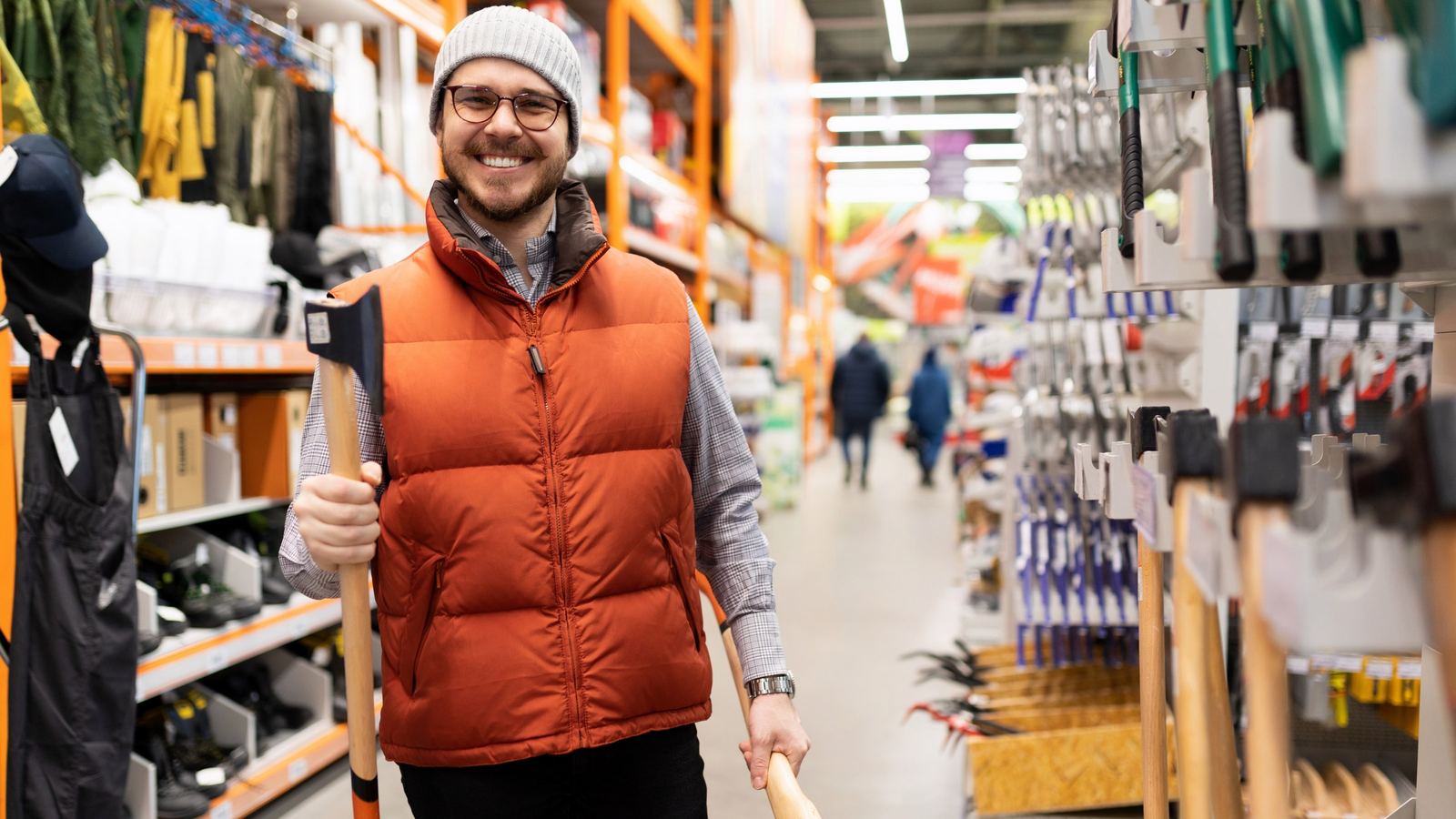 Smiling man in an orange vest and knit cap carrying an axe through a hardware store aisle