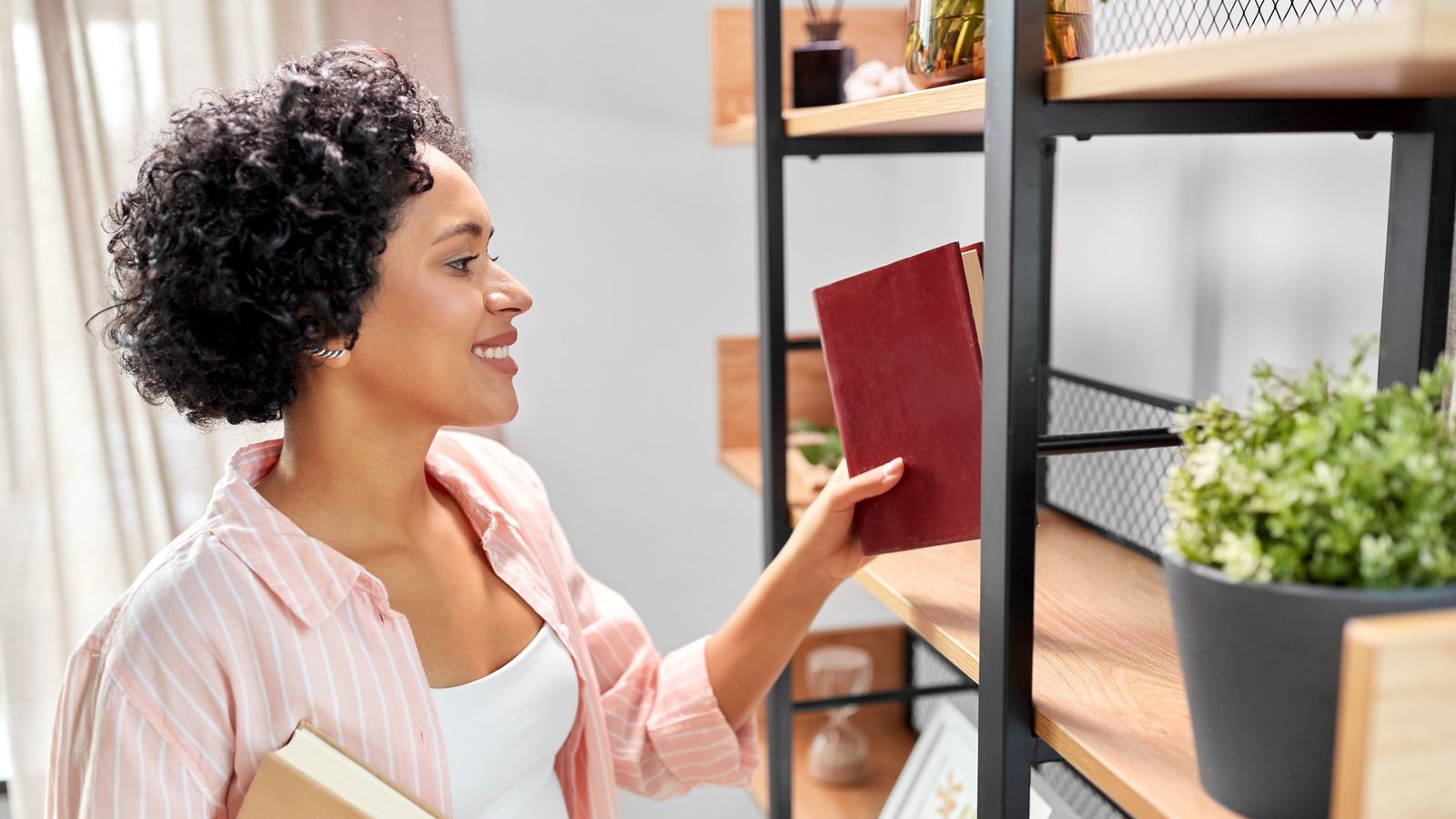 Smiling woman holding a book against her chest and reaching up to place a red book on a library shelf