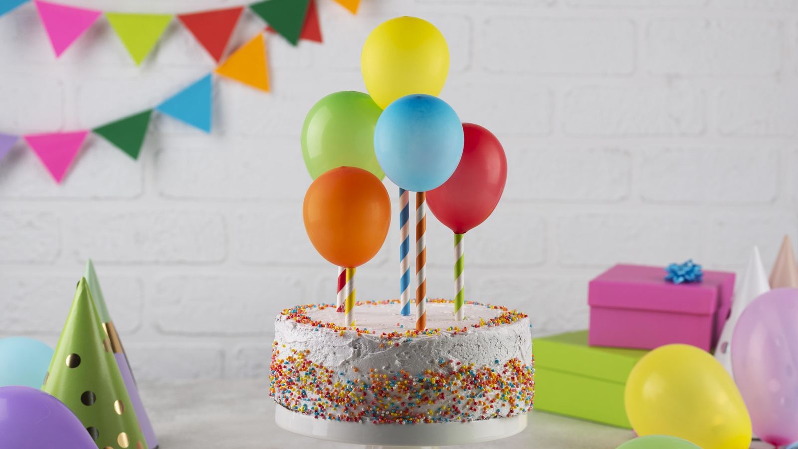 Birthday cake with colorful balloons, party hats and wrapped gifts on a table against a white brick wall