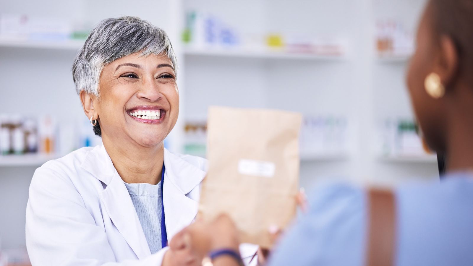 Smiling pharmacist in a white coat handing a paper pharmacy bag to a customer across the counter