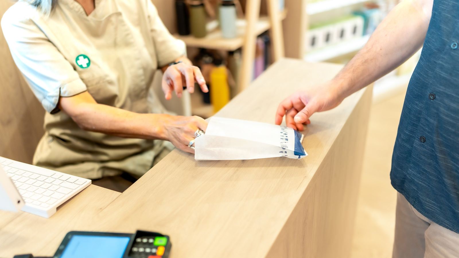 Store employee in a green apron at a wooden checkout counter handling a receipt for a customer, with a card reader beside them