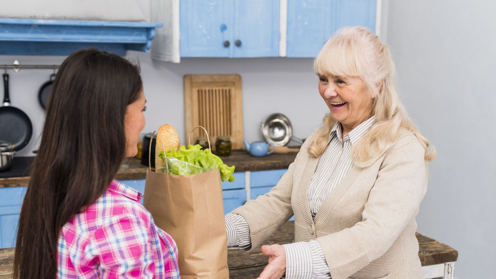 Young woman in a plaid shirt handing a paper bag of fresh vegetables to a smiling older woman in a kitchen
