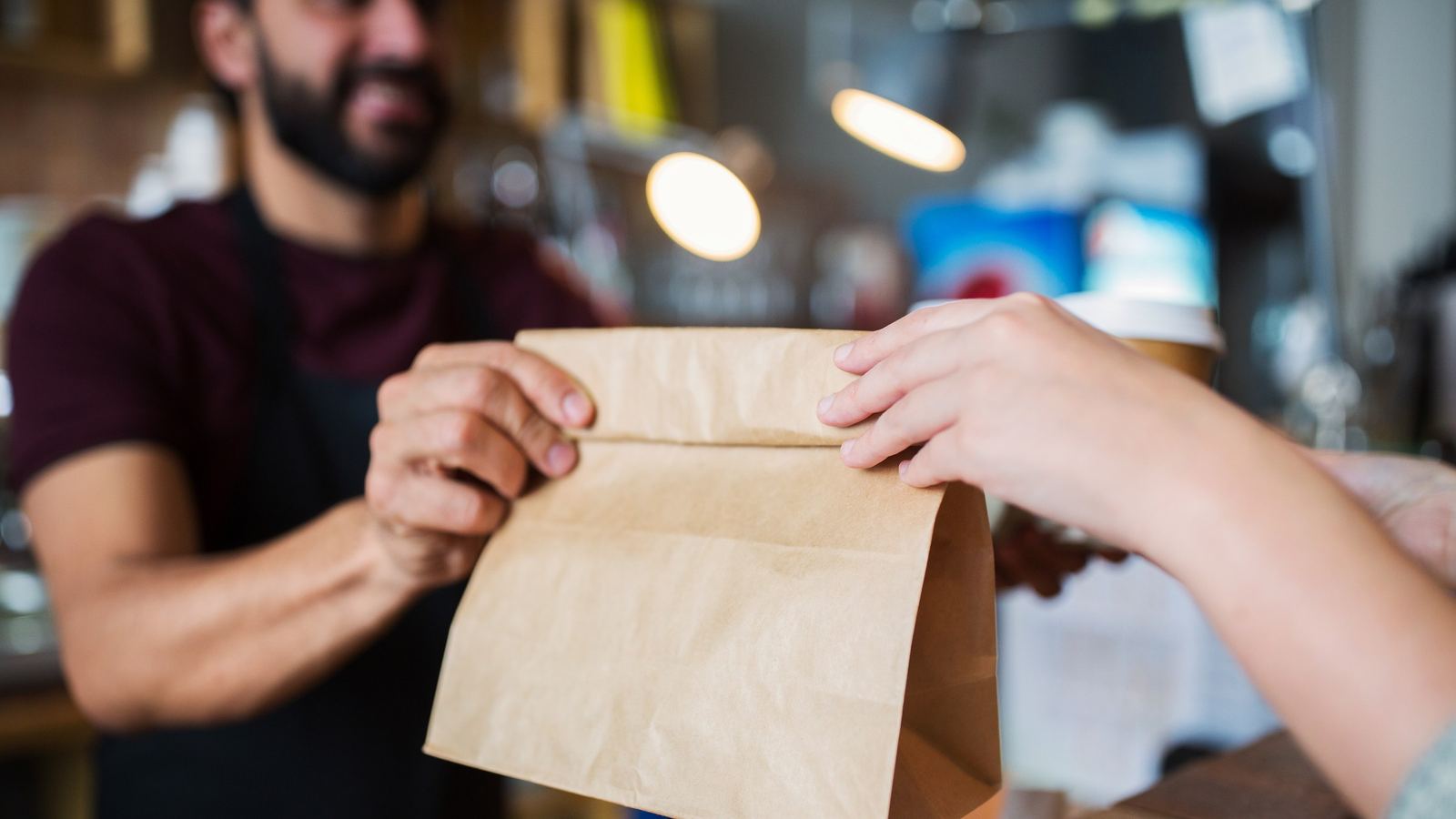 Man in an apron handing a brown paper takeout bag across a restaurant counter to a customer